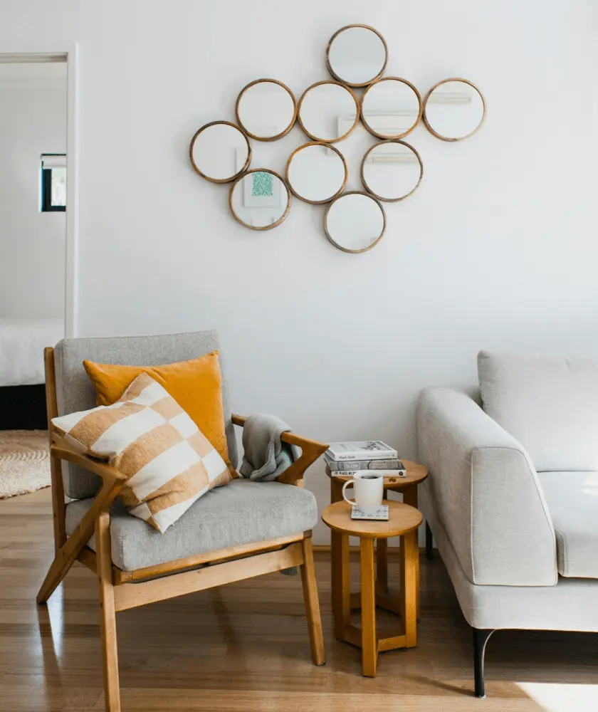 A bright living room features a light gray armchair with yellow and striped pillows, a light-colored sofa, and a wall adorned with a cluster of round, gold-framed mirrors.