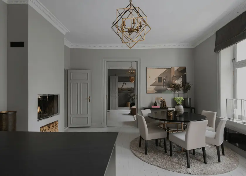 A gray dining room with a round black table, white chairs, and a geometric gold chandelier. To the left is a fireplace with stacked logs, and a doorway leads to another room.