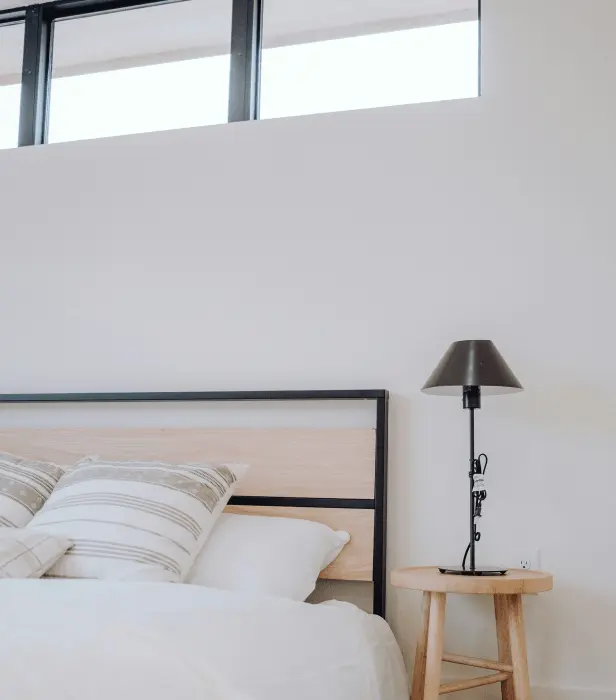 A modern bedroom with a white bed, wooden headboard, black lamp on a side table, and high windows